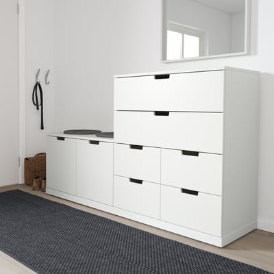 Moderne hvid NORDLI kommode i entreen, med gråt tæppe. Høj enhed med seks skuffer og sorte håndtag, placeret ved kroge og bænk.