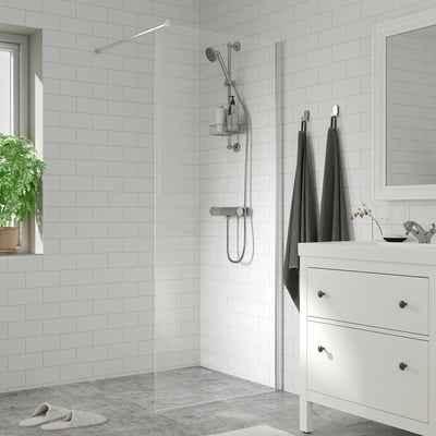 Moderne badeværelse med hvide fliser, glasbruser, kromarmaturer, grå håndklæder, lille håndvask, spejl og plante.