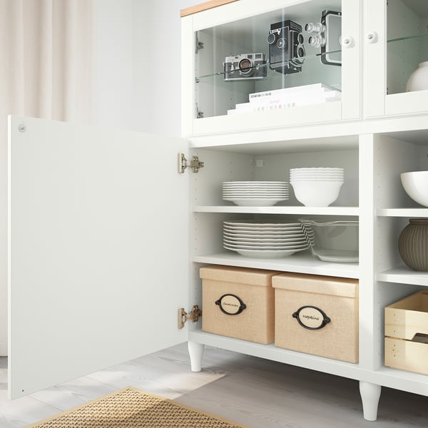 Hvidt skab med glasdør med vintage-kameraer og organiserede retter. Nederste hylder rummer beige opbevaringsbeholdere.