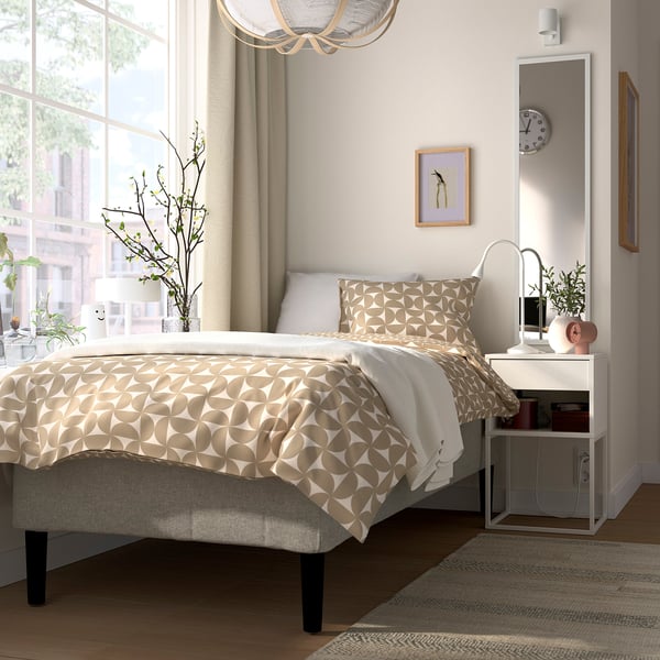 Hyggeligt soveværelse med beige RÅHOLMAR seng, natbord, ur, plante og stort vindue.