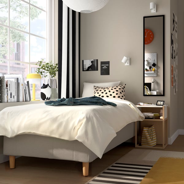 Moderne soveværelse med hvid RÅHOLMAR-seng, sort prikket pude, sideborde, bøger, plante, ur, taske, farverig kunst, stort vindue, mønstret tæppe.