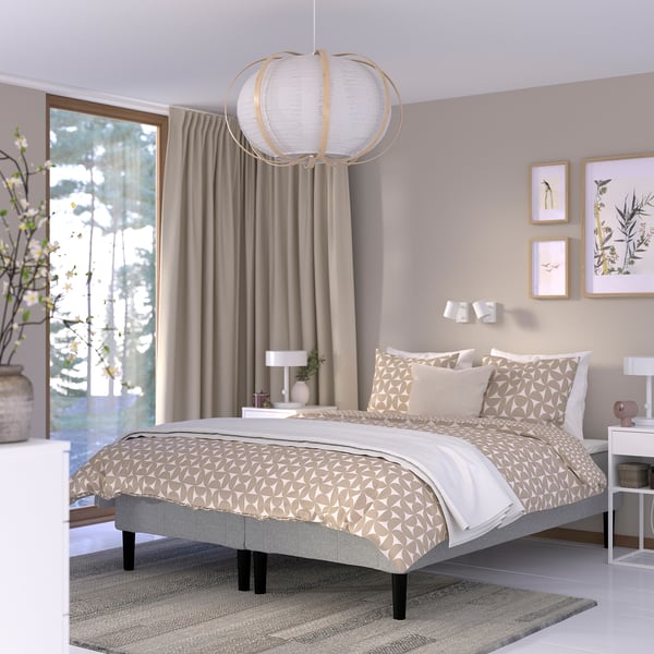 Moderne soveværelse med mønstret dyne, beige vægge, kunst, sfærisk lys, plante, vindue, natbord, lampe, neutrale farver.