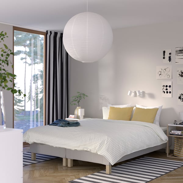 Soveværelse med stor hvid RENFJÄLLET seng, gule puder, sorte gardiner, stribet tæppe, hængende hvid lampe, plante og vægkunst.