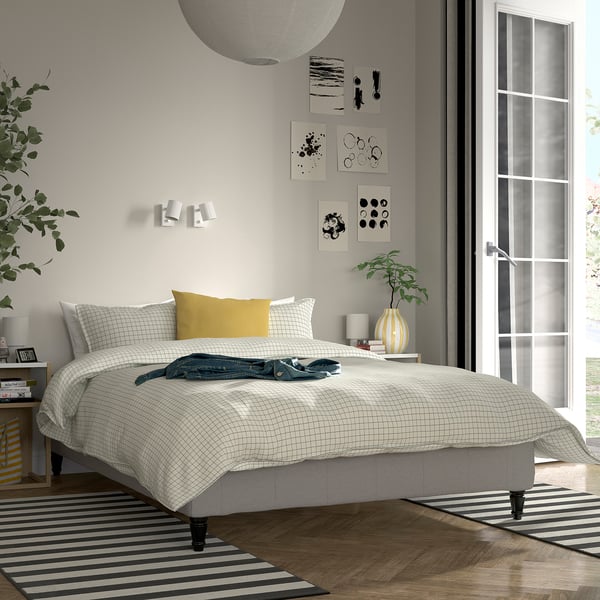 Moderne soveværelse med hvid RENFJÄLLET seng, gul pude, ternet opslag, vægkunst, natbord med lampe og plante, stribet tæppe, store vinduer.