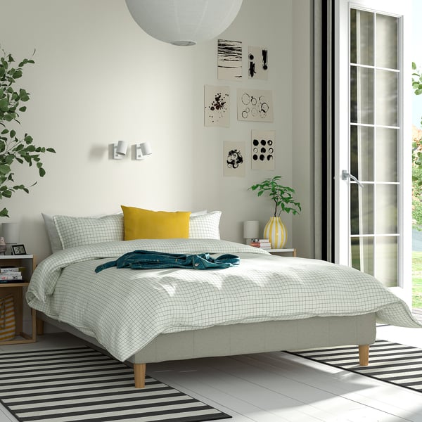 Moderne minimalistisk soveværelse med lav hvid seng, blåt tæppe, gul pude, stribet tæppe, planter og kunst. Sengen er større end natbordet.