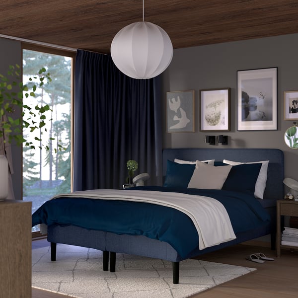Moderne soveværelse med mørkeblå seng, blødt hovedgærde, hvide puder, gråt sengetøj, trægulv, stort vindue, lampe, kunstværker, plante og natbord.