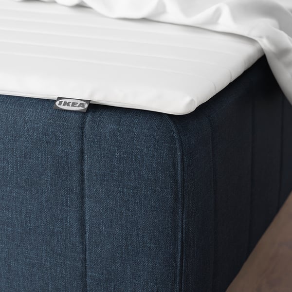 Close-up af et madrashjørne med mørkt betræk og hvid topper, mærket IKEA.