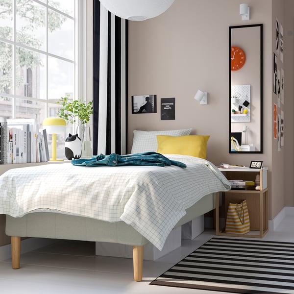 Moderne soveværelse med RENFJÄLLET-seng, gul pude, turkis tæppe. Ved siden af sengen, bord med bøger, ur. Stribet gulv, stort spejl, levende kunst.