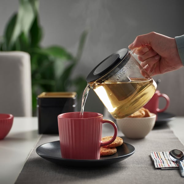 Person hælder te fra en glastekande med en infusionsenhed i et rødt krus, siddende på en sort underkop ved siden af småkager og en ske.