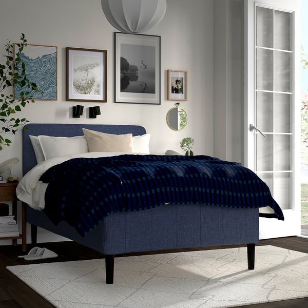 Et moderne soveværelse med en blå RISHÖJDEN-seng som midtpunkt, prydet med marineblåt sengetøj, flankeret af et hvidt tæppe og omgivet af minimalistisk indretning.