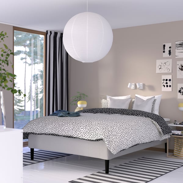 Moderne soveværelse, grå og hvid indretning, stor seng, pendel, stribet tæppe, vindue med mørke gardiner, minimalistisk natbord med lampe og plante.
