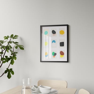 Indrammet abstrakt kunst hænger på væggen i en spisestue, flankeret af en træstol og en potteplante.