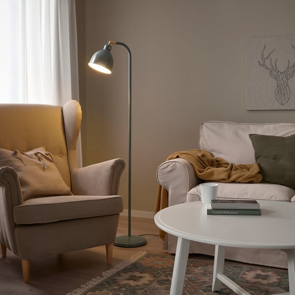 Hyggelig stue med RÖDFLIK lampe, beige stol, sofa, tæppe, bøger og tæppe. Lampen står højt nær vinduet og kaster varmt lys.