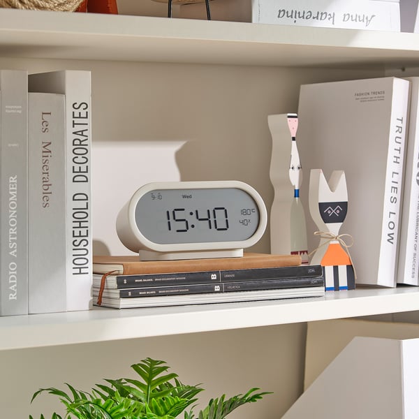 Bøger, ur, dekoration, plante på hylden. Uret viser tid, temperatur og fugtighed.