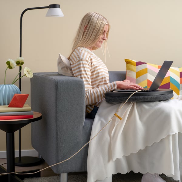 Kvinde i stribet skjorte arbejder på laptop ved bordet med lampe og blomster.