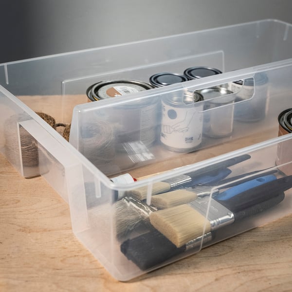 Gennemsigtig plastikorganisering med håndtag, der holder pensler, små glas og snore, ideel til kunst og håndværk.