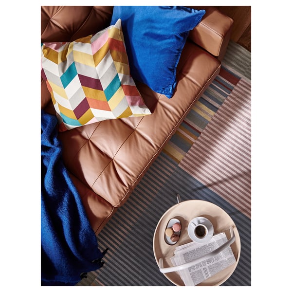 Stue med brun SANELA pude, farverig plaid, blåt tæppe, kaffekop, fjernbetjening og magasin.