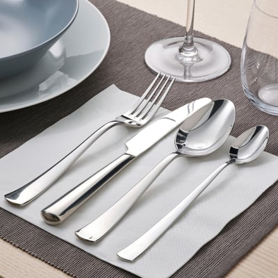 Bestik i poleret rustfrit stål sat på en serviet, inklusive en kniv, gaffel og ske, arrangeret på et bord med tallerkener og glas.