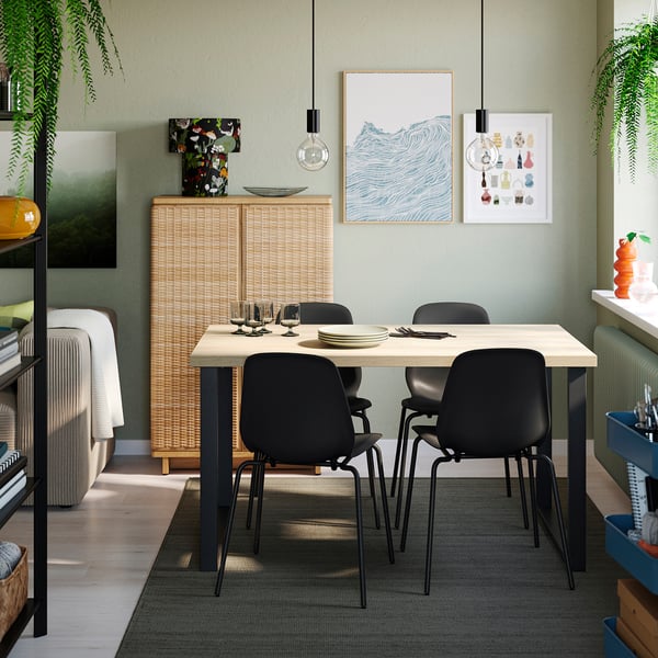 Moderne spiseplads med SKÅLSTA bord og sorte stole. Lyst træbord, mørke metalben. Lys kunst, planter, kurveskab. Hyggelige, afslappede omgivelser.