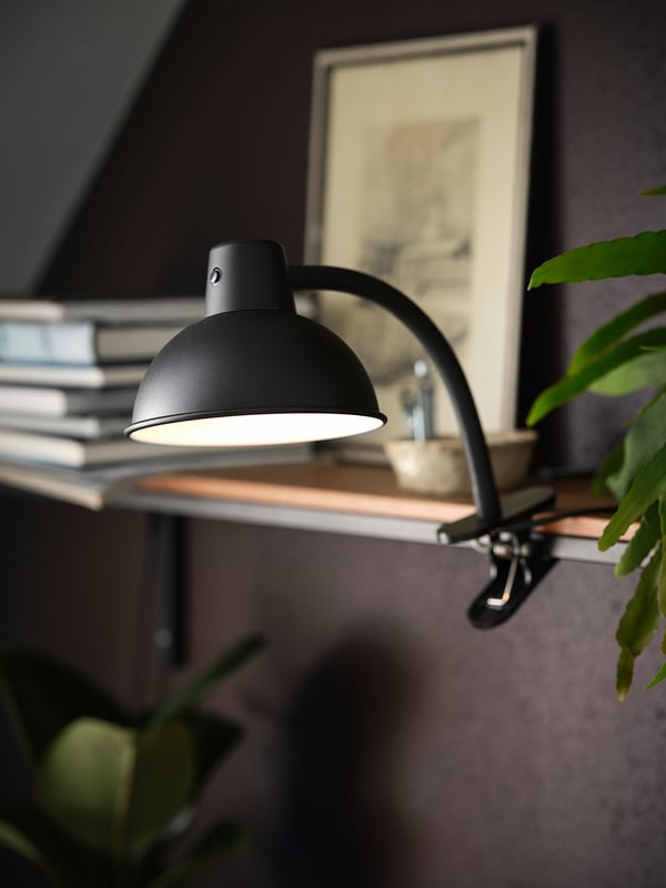 En moderne sort SKURUP-lampe med en buet arm klemmes fast på en hylde og kaster lys på en plante og bøger.