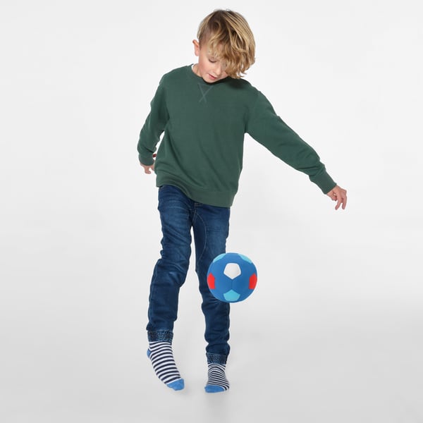 Barn balancerer farverig bold på knæet, iført grøn sweater, jeans og stribede sokker.