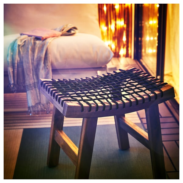 Brun akacietræ udendørs skammel med vævet sæde og ben, ved siden af seng med tæppe.