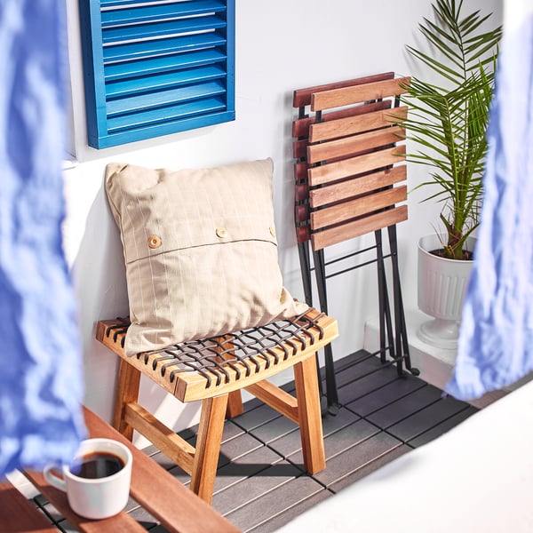 Altanscene med STACKHOLMEN træskammel, beige pude, kaffekrus, potteplante, blå skodder og lyseblå gardiner.