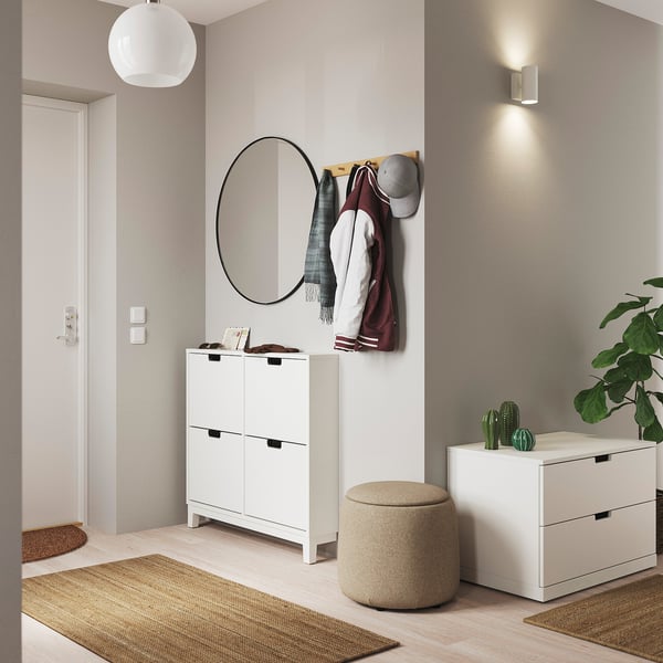 Moderne entré med hvide møbler: rundt spejl, skoskab og skammel. Planter og hatte på kroge. Lyse trægulve, beige tæpper.