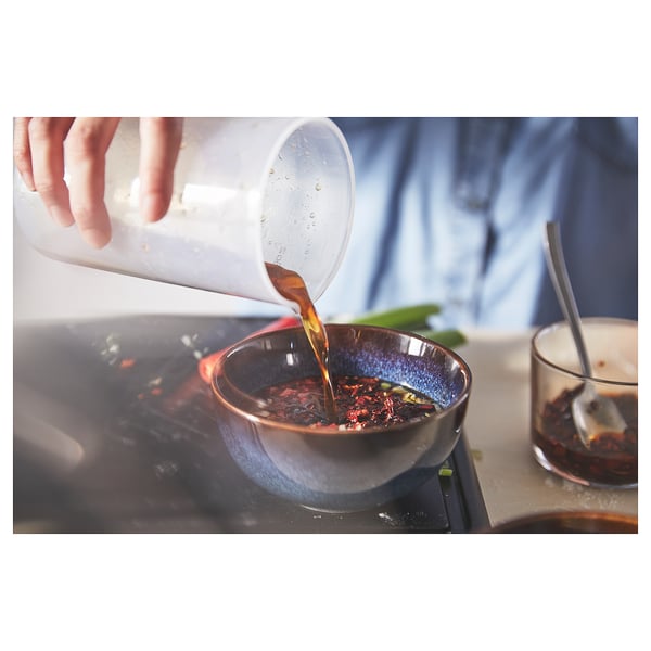 Håndhælder væske fra en klar plastikkande i en blå glasskål på et komfur. Væsken er ravfarvet.