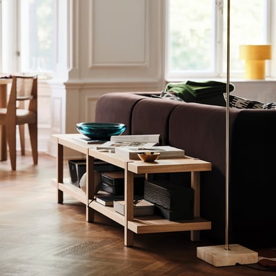 STOCKHOLM 2025 bænk i lyst træ, nær mørkebrun sofa. Rummer magasiner, skåle og bøger. Forenklet, moderne design.