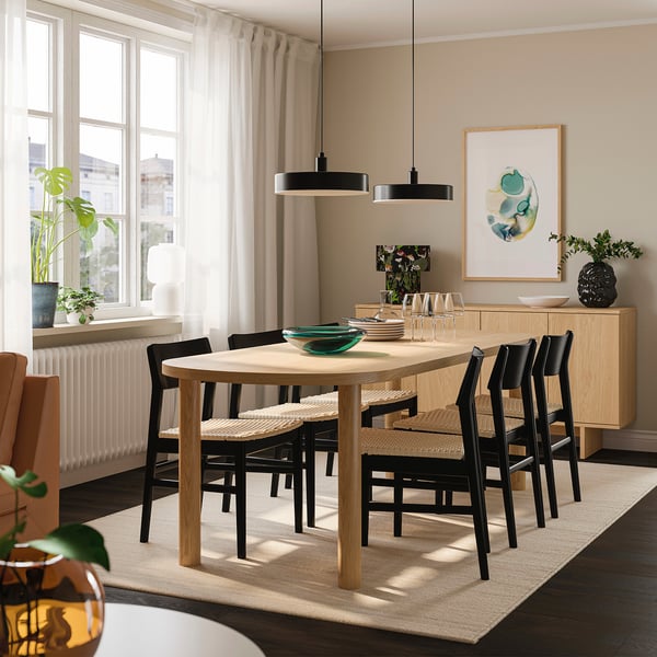 Moderne spisestue med lyst træbord, sorte stole, stort vindue, grøn plante og minimalistisk indretning.