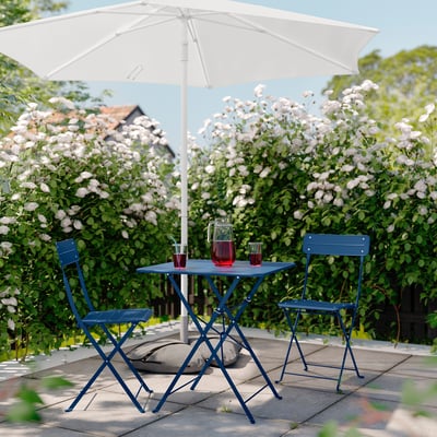 Blå metalterrassesæt med bord og stole under en hvid parasol, beliggende i en frodig have.