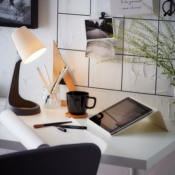 Moderne arbejdsplads med hvidt skrivebord, sort krus, tablet, lampe, kuglepenne og plante.