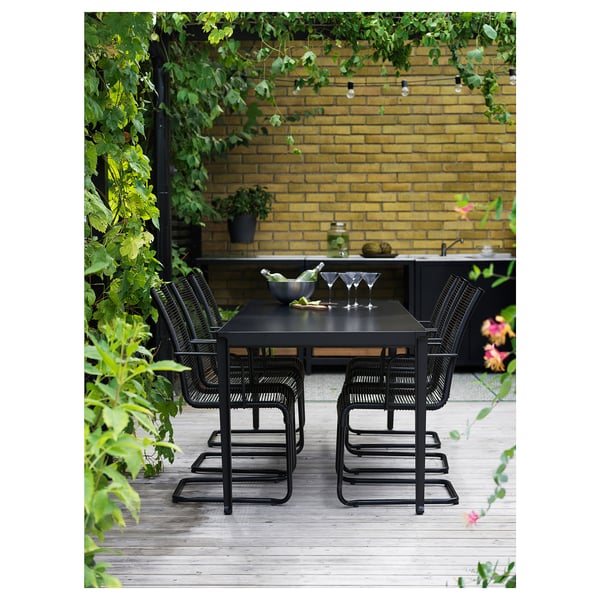 Moderne udendørs spisegruppe med sorte stole og bord på en træterrasse med en gul murstensvæg i baggrunden og frodige grønne områder.