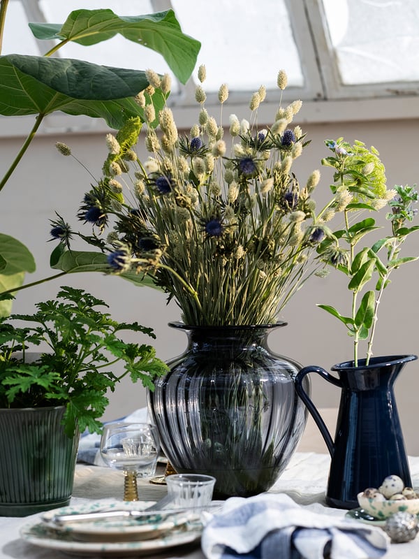 Glasvase med tørret lavendel og blå blomster, ved siden af en blå kande på bordet med grønne planter og tallerkener.