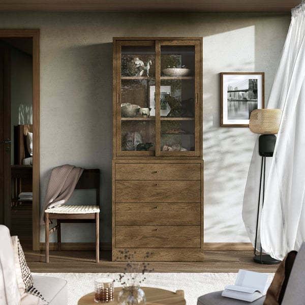 Hyggeligt værelse med TONSTAD opbevaring. Træskab, stort, med glaslåger og skuffer. Moderne, brune og beige toner. Gulvlampe til højre, stol til venstre.