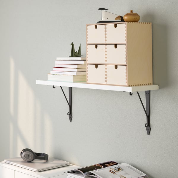 En væghængt hylde med skuffer med bøger og en dekorativ genstand. Nedenfor er der et skrivebord med hovedtelefoner, en bærbar computer og magasiner.