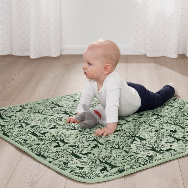 Baby på måtte med skovtema, liggende på maven og udforske plyslegetøj.