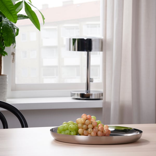 Metalbakke med druer på træbord nær vindue, lampe i baggrunden.