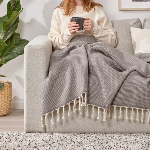 Person, der sidder på en grå sofa, pakket ind i en grå uldplaid, med frynser i hånden, med et sort krus i hånden.