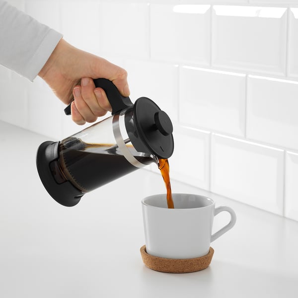 Håndhældning af kaffe fra en sort stempelkande i et hvidt krus på en korkunderlag mod en væg med hvide fliser.