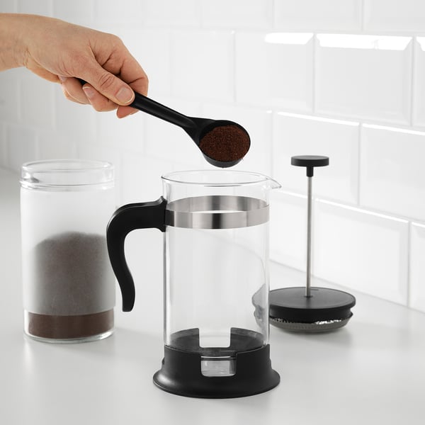 En hånd hælder kaffegrums i en fransk presse af klart glas, med et stempel klar til at brygge på en hvid bordplade.