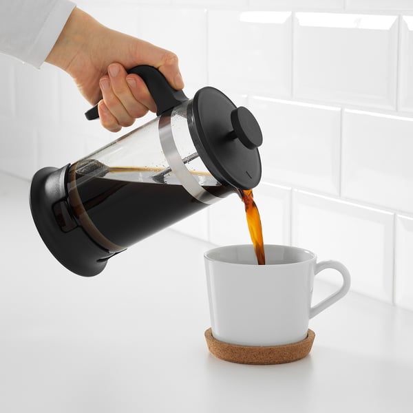 Person hælder frisklavet kaffe fra en fransk presse i et hvidt krus på en korkunderlag.