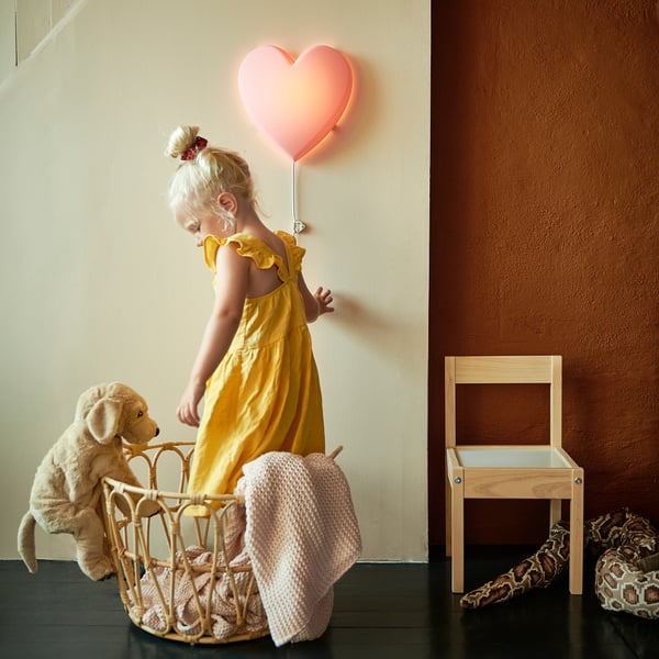 Lille pige i gul kjole skubber klapvogn med bamse. Hjerteformet lampe på væg, træstol, slangeskindslegetøj.