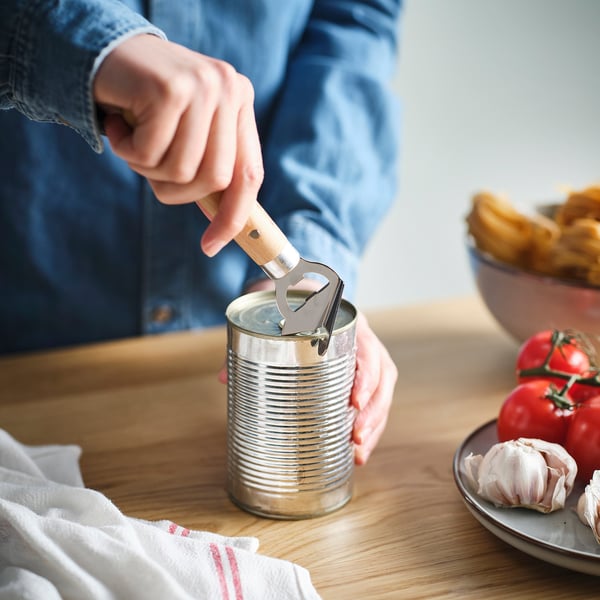 Person åbner dåse ved hjælp af VARDAGEN flaske/dåseåbner på træbord, med tomater og hvidløg i nærheden.