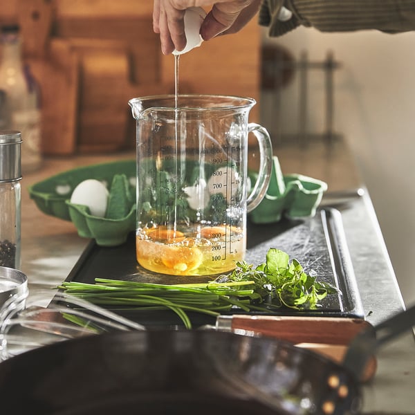 Person blander væske i en klar målekande med ingredienser og urter på et køkkenbord.