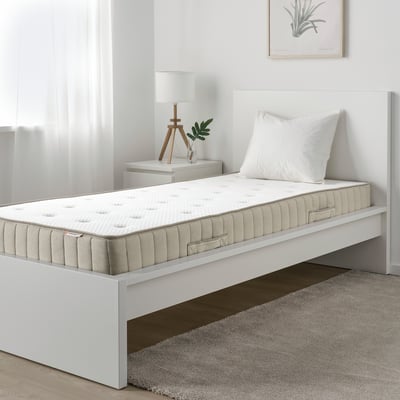 Moderne minimalistisk soveværelse, hvid enkeltseng med madras, hvid pude, lille hvidt natbord med lampe og plante.