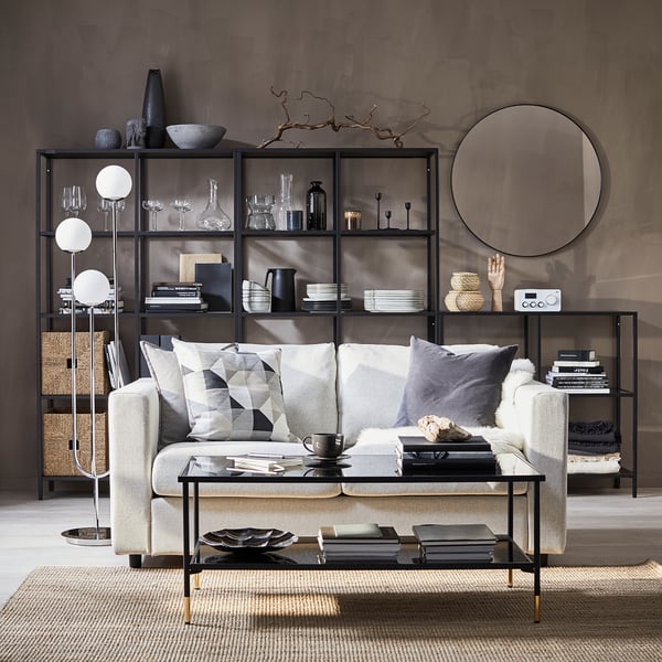 Moderne stue en grå VIMLE sofa med geometriske puder, et sofabord i glas og en bogreol i sort metal fyldt med dekorative genstande.