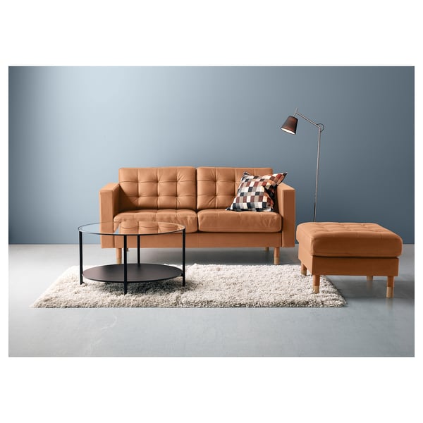 Moderne stueopsætning med orange sofa, matchende fodstøtte, sofabord i glas og gulvlampe mod blå væg.