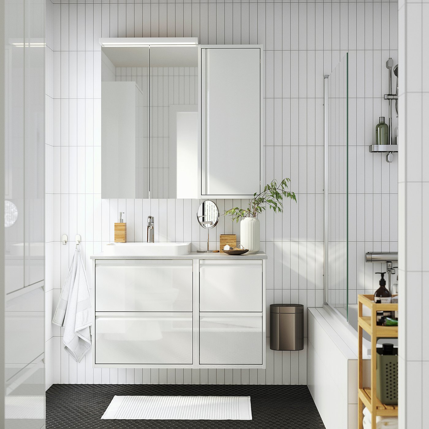 Modern white bathroom with tall ÄNGSJÖN cabinet, sink, mirror, black bin, towels, plant, and cart.